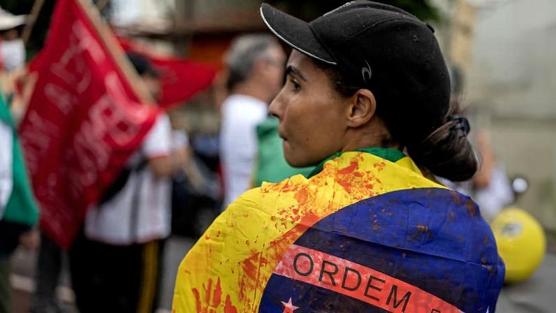 Pessoa de perfil usa boné preto e está envolta em bandeira do Brasil com manchas vermelhas. Ao fundo, outras pessoas e bandeira vermelha desfocadas. Bandeira brasileira manchada de vermelho é exibida por manifestante durante ato após operação com recorde de mortes na Penha e no Alemão, no Rio