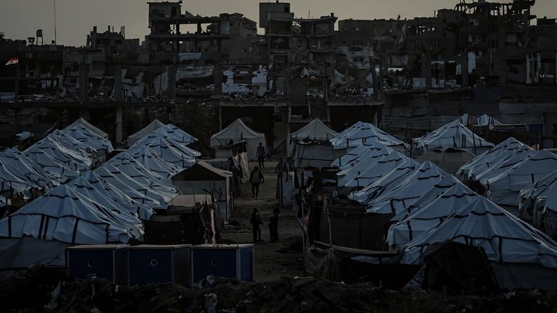 Campo de refugiados com fileiras de tendas brancas alinhadas em terreno aberto. Pessoas caminham entre as tendas. Ao fundo, prédios severamente danificados e destruídos, com estruturas expostas e paredes desabadas.