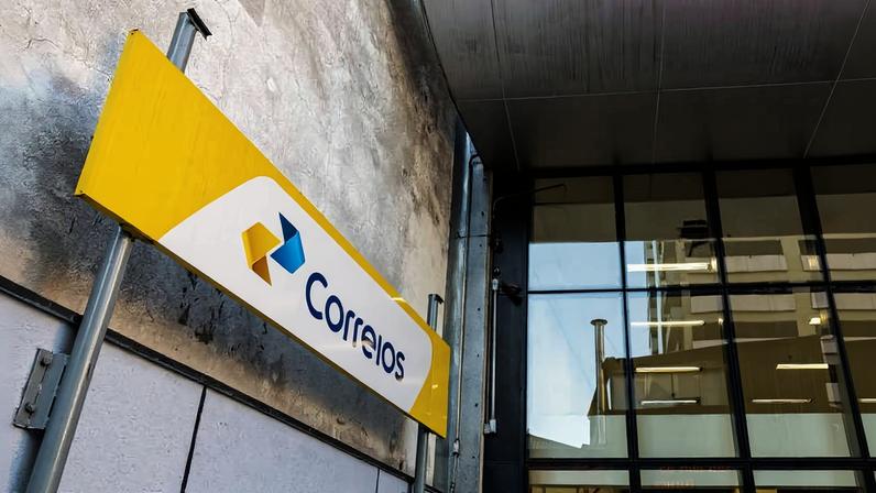 Entrada de agência dos Correios com placa amarela e branca exibindo o logotipo e nome da empresa. Fachada moderna com paredes de vidro e estrutura metálica.