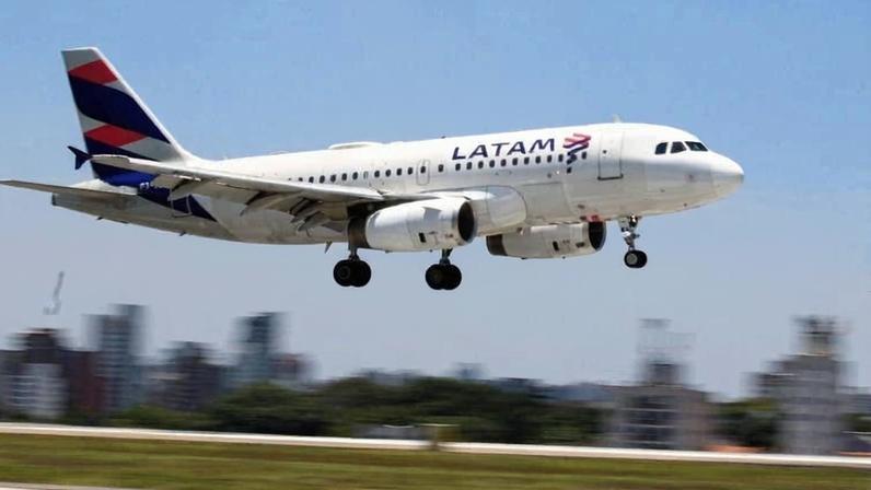 Avião comercial da LATAM com trem de pouso abaixado voa baixo sobre pista de aeroporto, com prédios urbanos desfocados ao fundo sob céu azul com poucas nuvens.