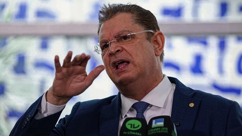 Homem de terno azul e gravata azul fala gesticulando com a mão direita levantada. Ele está diante de microfones com logos de canais de TV, em ambiente interno com fundo desfocado.