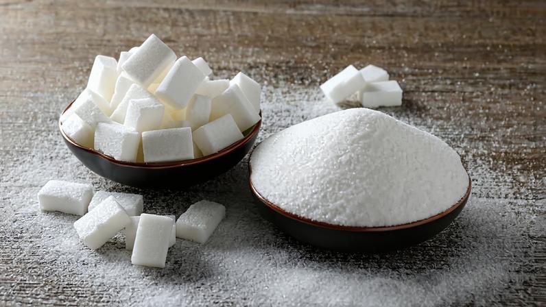 Duas tigelas pequenas sobre uma superfície de madeira clara, uma contendo cubos de açúcar branco empilhados e outra cheia de açúcar granulado branco, com açúcar espalhado ao redor.