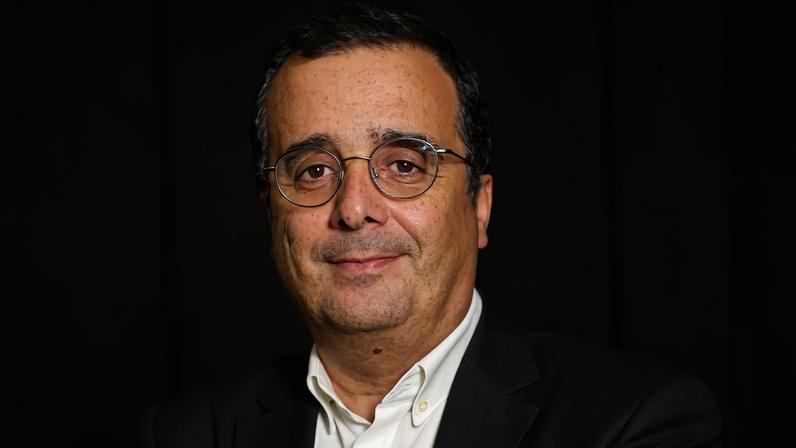 Homem de meia-idade com óculos e cabelo curto, veste camisa branca e blazer preto, está com os braços cruzados em frente a logo vermelha da ESPN em parede branca. Ao fundo, monitores e pessoas trabalhando em ambiente de redação.