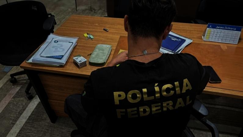 Agentes da Polícia Federal visto de costas sentado em cadeira diante de mesa de madeira com documentos, carteira e objetos pequenos sobre a superfície.
