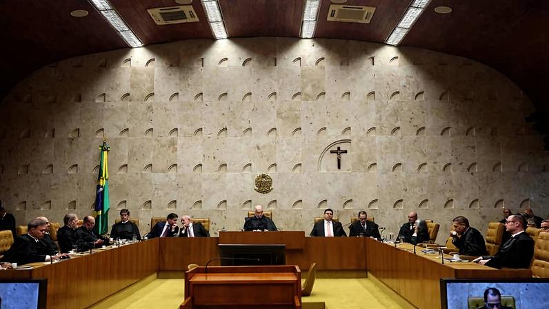 Ministros sentados em semicírculo no plenário do Supremo Tribunal Federal, com parede de pedra ao fundo, crucifixo e bandeira do Brasil à esquerda.