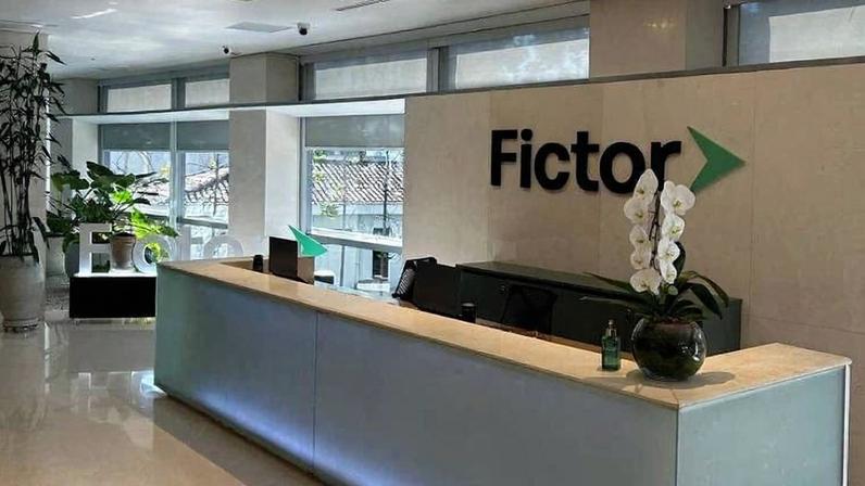 Balcão de recepção iluminado com luz azul em ambiente corporativo claro. Parede ao fundo exibe logo 'Fictor' em preto com seta verde. Plantas decorativas estão ao lado e sobre o balcão.