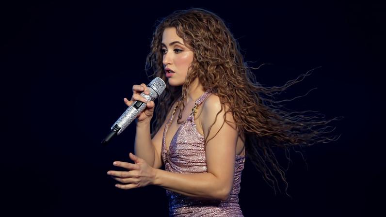 Uma artista está se apresentando em um palco, segurando um microfone. Ela usa um vestido brilhante de cor rosa e tem cabelos longos e cacheados. O fundo é escuro, com luzes amarelas visíveis ao redor, sugerindo um ambiente de show.