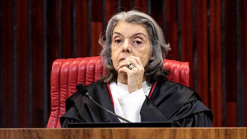 Ministra do Supremo Tribunal Federal sentada em cadeira vermelha, vestindo toga preta com detalhes vermelhos, com mão próxima ao rosto, em ambiente formal com painel de madeira ao fundo.