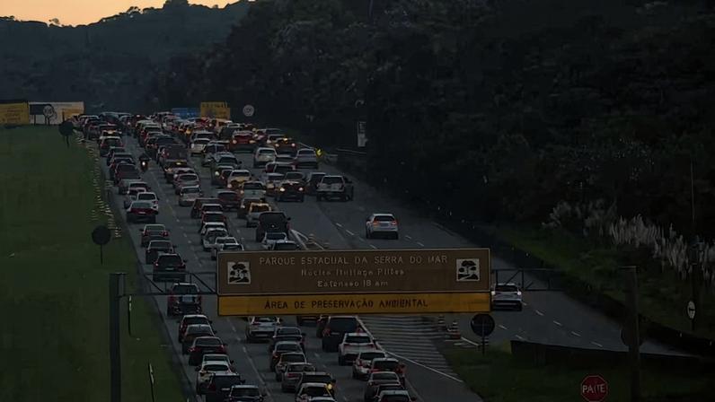 Congestionamento de veículos em múltiplas faixas de uma rodovia cercada por vegetação. Placa indica entrada para o Parque Estadual da Serra do Mar e área de preservação ambiental. Sinal de pare visível à direita.