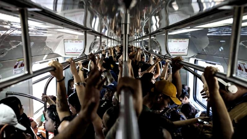 Interior de vagão de metrô cheio de passageiros em pé segurando barras de apoio metálicas no teto. Pessoas estão próximas umas das outras, algumas usando bonés e mochilas. Luz natural entra pelas janelas laterais.