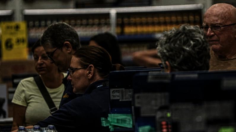 Cinco pessoas próximas a caixas registradoras em supermercado. Funcionária de uniforme azul atende clientes, entre eles um homem idoso e três adultos, em ambiente interno com prateleiras ao fundo.