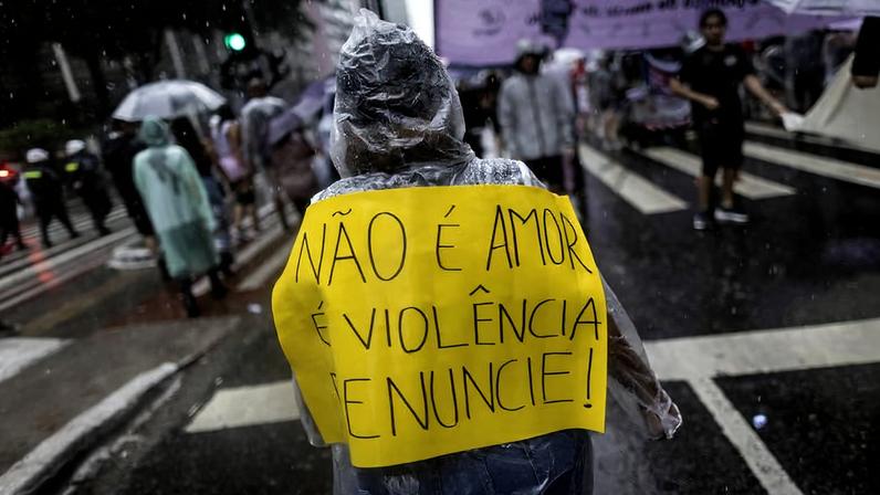 Pessoa vestindo capa de chuva transparente segura cartaz amarelo nas costas com a frase 'NÃO É AMOR É VIOLÊNCIA DENUNCIE!' durante protesto em rua molhada. Outras pessoas com capas de chuva e guarda-chuvas aparecem ao fundo em dia chuvoso.
