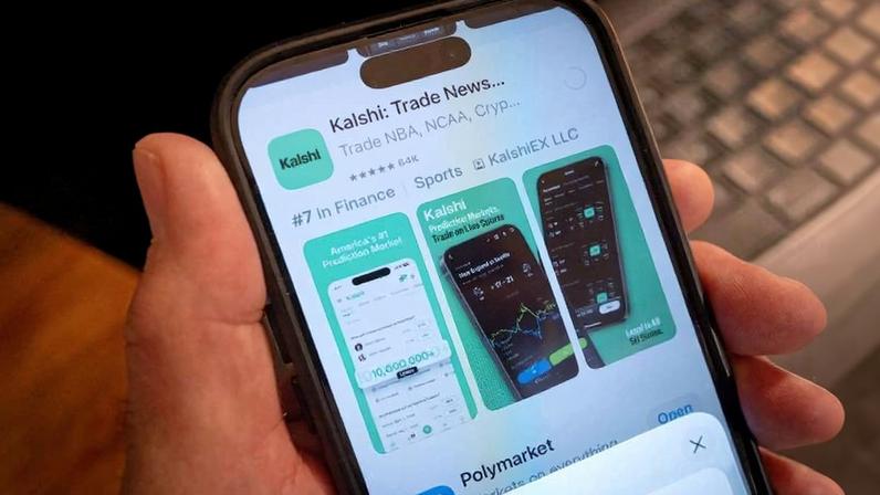 Mão segurando smartphone com a tela exibindo a página do aplicativo Kalshi Trade News na loja digital. A tela mostra avaliações, categoria Financeira, e imagens de pré-visualização do app com gráficos e dados financeiros. Fundo desfocado com teclado ao fundo.