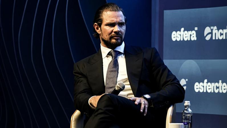 Homem de cabelo preso e barba, vestido com terno escuro e gravata, sentado em cadeira branca segurando microfone. Fundo azul com logotipo 'esfera' repetido em painel.