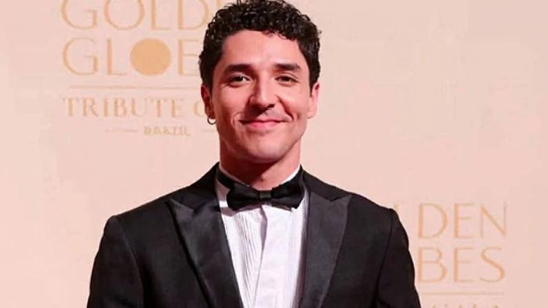 Homem jovem com cabelo curto e encaracolado veste smoking preto com gravata borboleta preta e camisa branca, posando com uma mão no bolso em fundo claro com texto desfocado relacionado ao Golden Globes.