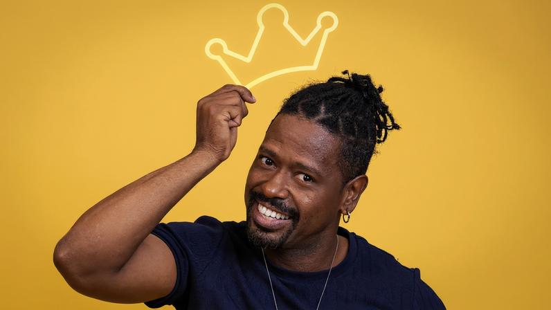 Homem de pele escura com cabelo preso em dreadlocks sorri enquanto segura uma coroa desenhada em vermelho acima da cabeça, com fundo amarelo.