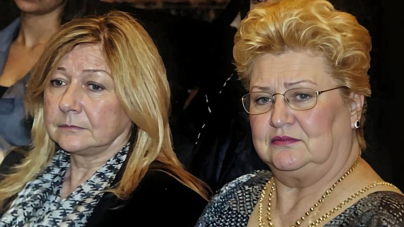 Duas mulheres sentadas lado a lado em ambiente interno. A mulher à esquerda tem cabelo loiro claro e veste casaco preto com lenço estampado. A mulher à direita tem cabelo curto e loiro, usa óculos, colar dourado e blusa cinza.