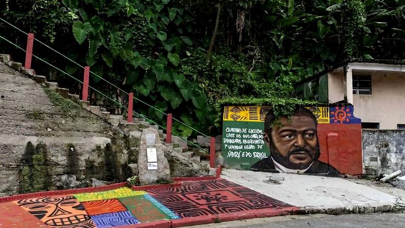 Mural colorido retrata o rosto de um homem negro com expressão séria, ao lado de texto em português. O mural está pintado em uma parede próxima a escadaria de concreto com corrimão vermelho, rodeada por vegetação densa. O chão à frente do mural também é decorado com padrões geométricos coloridos.