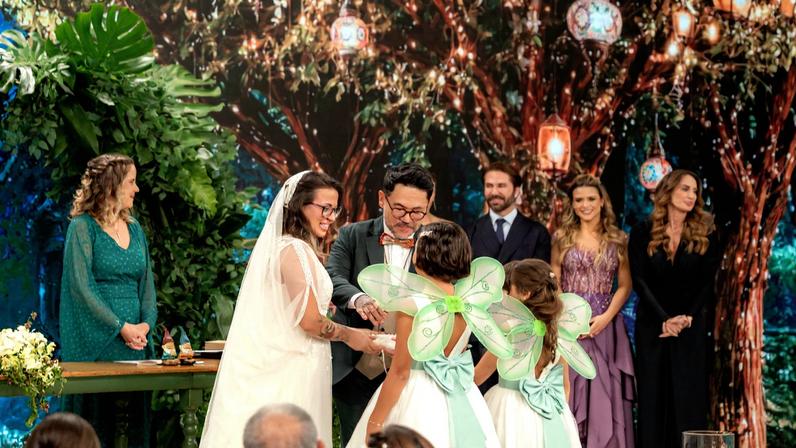 Noivos trocam alianças diante de três daminhas vestidas de branco com asas verdes. Ao fundo, quatro pessoas observam, e decoração inclui folhagens e luzes pendentes.