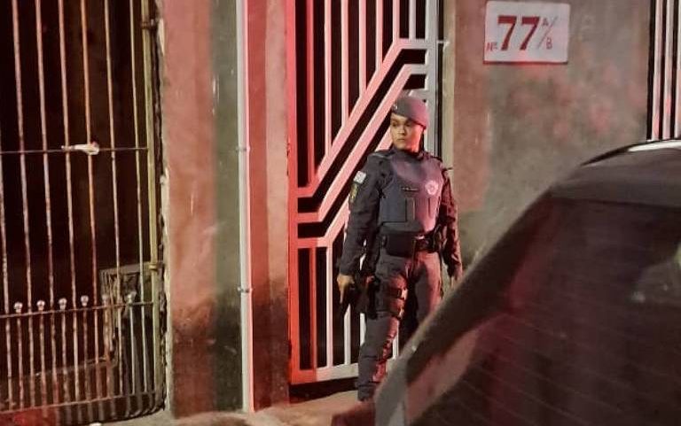Policial armado está parado em frente a uma porta de grade vermelha numerada como 77, em uma rua à noite. Veículo da polícia está estacionado próximo, com parte da palavra 'POLÍCIA' visível na lataria.