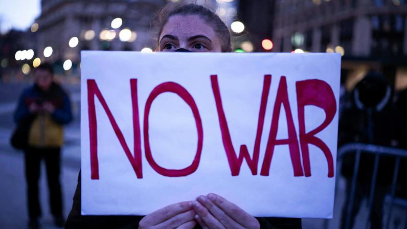 Pessoa segura cartaz branco com letras vermelhas 'NO WAR' em local público ao entardecer. Luzes urbanas desfocadas ao fundo.