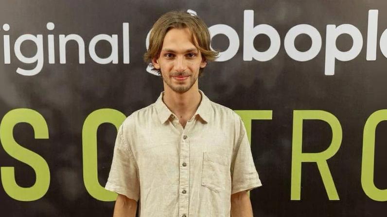 Homem jovem de cabelos médios loiros usa camisa bege de manga curta e calça clara, posando em tapete vermelho diante de painel preto com logo da Globoplay e texto em verde e branco.