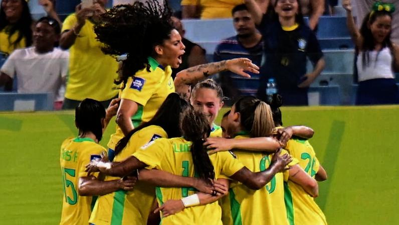 Seis jogadoras da seleção brasileira feminina de futebol se abraçam em comemoração, com uma delas saltando animada. Ao fundo, torcedores vibram nas arquibancadas.