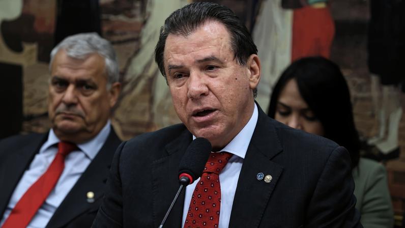 Homem de meia-idade com terno cinza e gravata vermelha fala em microfone em ambiente formal. Ao fundo, duas pessoas sentadas, uma delas com gravata vermelha.