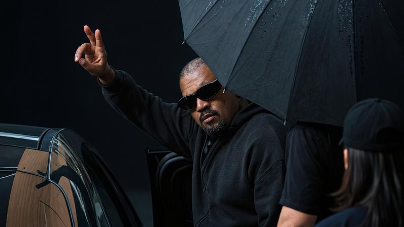 Homem com moletom preto e óculos escuros levanta a mão fazendo sinal de paz enquanto está ao lado de um carro preto. Uma pessoa segura um guarda-chuva preto sobre ele, e outra pessoa está parcialmente visível de costas.