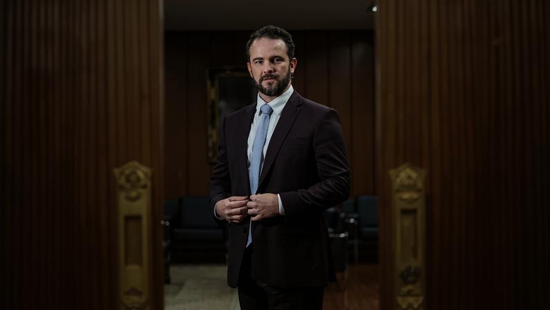 Homem de terno vinho e gravata azul claro está em pé em sala com paredes de madeira e piso de madeira. Ele está centralizado entre duas portas de madeira parcialmente abertas com maçanetas douradas. Ao fundo, há poltronas pretas e um quadro na parede.