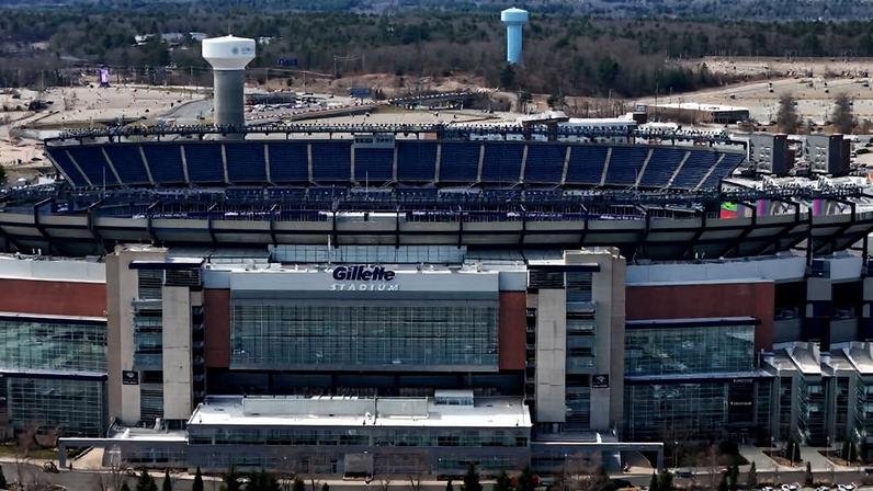 Vista aérea do estádio Gillette com arquibancadas vazias e área de estacionamento parcialmente ocupada ao redor. Ao fundo, áreas de vegetação e duas torres de água visíveis.