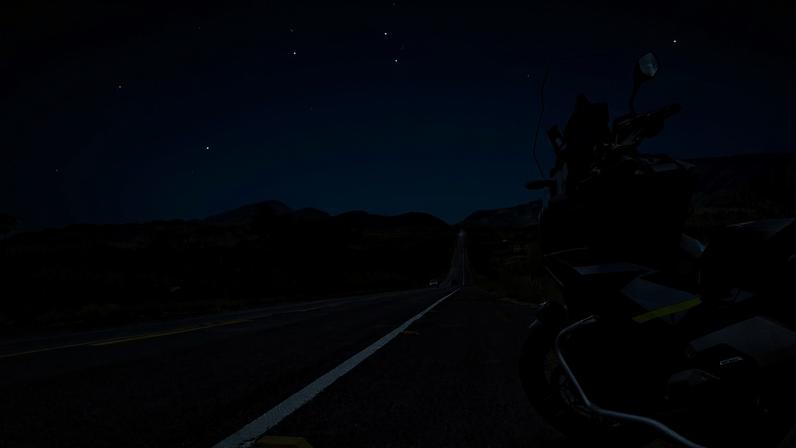 Duas motocicletas estão estacionadas lado a lado na margem de uma estrada asfaltada com faixa amarela dupla. Ao fundo, há um campo de cultivo dourado sob céu azul claro e sem nuvens.