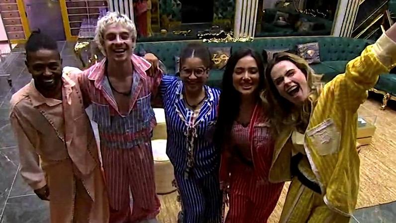 Cinco pessoas posam juntas sorrindo em sala com sofá verde ao fundo. Elas usam pijamas listrados em tons de rosa, azul, vermelho e amarelo, com ambiente interno iluminado.