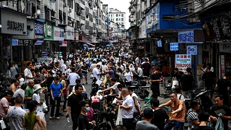 Rua estreita em área urbana densamente povoada com centenas de pessoas caminhando e interagindo entre lojas e edifícios residenciais. Muitas pessoas estão próximas a motocicletas e bicicletas estacionadas, com fachadas comerciais visíveis e letreiros em chinês ao longo da via.