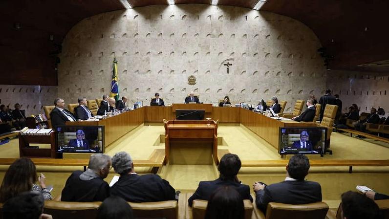 Ministros sentados em semicírculo no plenário do Supremo Tribunal Federal, com público em bancos à frente. Parede de fundo tem crucifixo e brasão da República, e bandeira do Brasil está à esquerda.