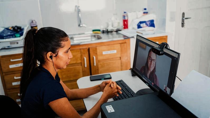 Mulher sentada em frente a computador em cozinha, participando de videoconferência com outra mulher visível na tela. Ela usa fones de ouvido e está concentrada na conversa. Ao fundo, armários de madeira, pia e utensílios de cozinha.