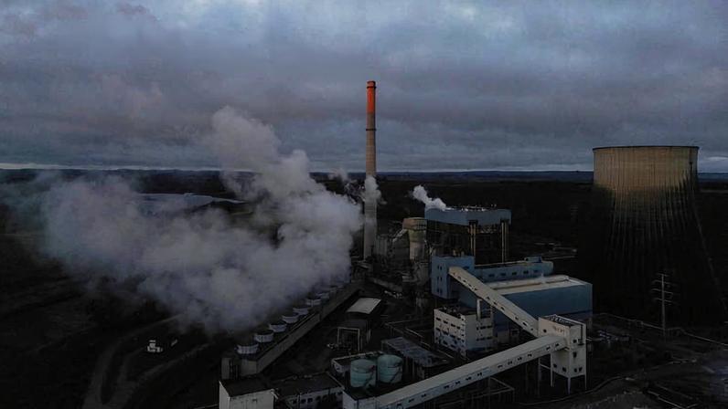 Usina termelétrica com chaminé liberando fumaça alaranjada pelo pôr do sol. Estruturas industriais e torre de resfriamento visíveis sob céu nublado.