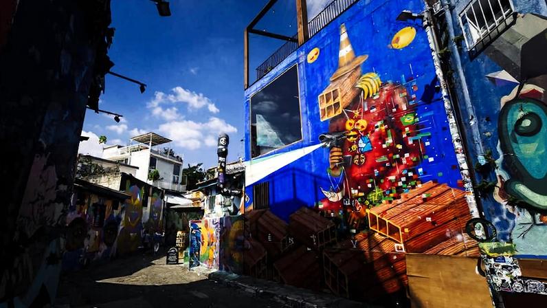 Grafite vibrante cobre parede azul em viela estreita, mostrando figura abstrata com elementos geométricos e cores vivas. Céu azul com poucas nuvens ao fundo e outras paredes com arte urbana visível na sombra.