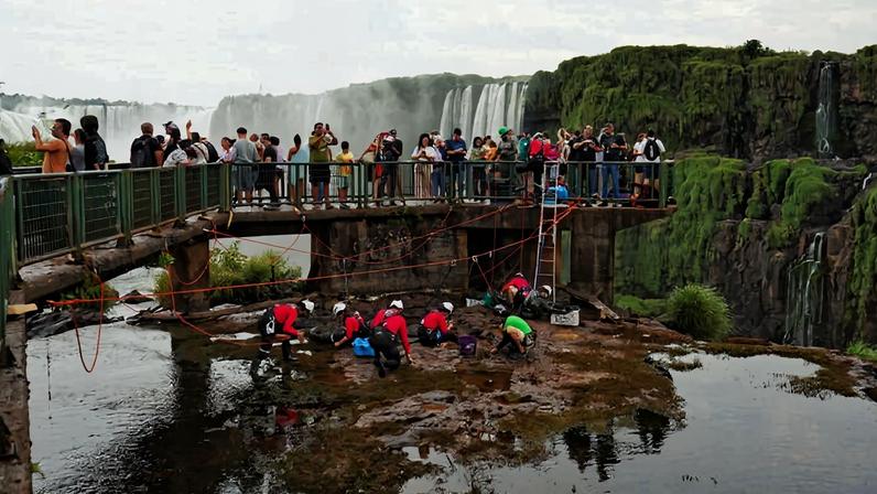 Equipe de trabalhadores com capacetes vermelhos realiza manutenção na base de uma passarela sobre rio próximo às Cataratas do Iguaçu. Turistas observam a atividade na passarela acima, com quedas d'água e vegetação ao fundo.