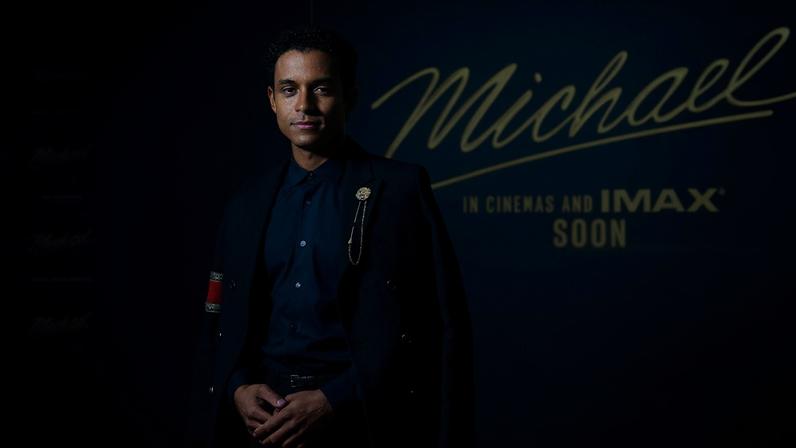 Homem jovem veste terno preto com camisa escura, posando em frente a painel azul escuro com texto dourado 'Michael' e 'In cinemas and IMAX soon'.
