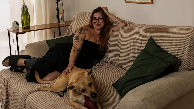 Mulher de cabelos longos e óculos está deitada em sofá coberto por manta, acariciando cachorro grande deitado ao seu lado. Ao lado do sofá, manequim masculino sem roupas usa chapéu marrom. Na parede, três quadros com textos e imagens em vermelho e branco. Ambiente iluminado por luz natural de janela à esquerda.