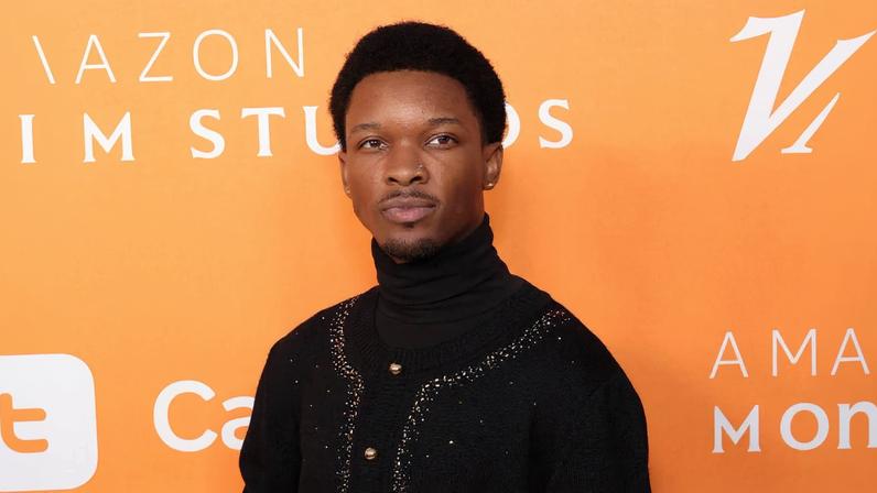 Homem jovem com roupa preta elegante, incluindo luvas e detalhes dourados, posa em frente a fundo laranja com logos da Amazon Studios e Variety.