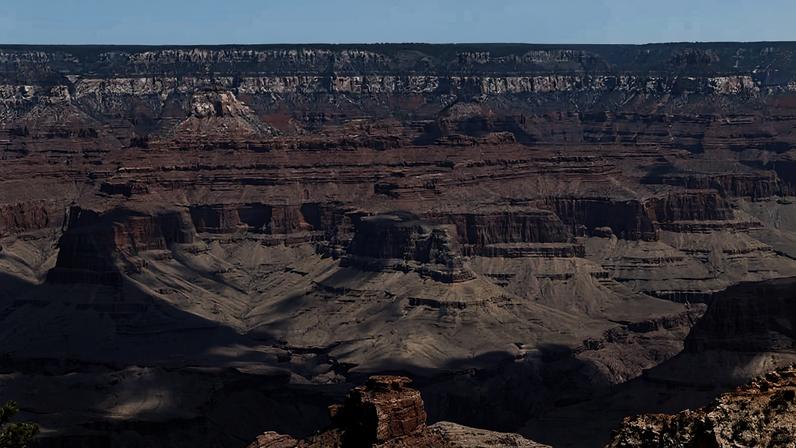 Formações rochosas e camadas geológicas do Grand Canyon sob céu azul com poucas nuvens. Sombras destacam a profundidade do cânion.