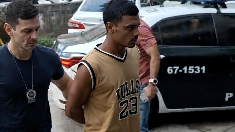 Homem de camiseta regata marrom e bermuda clara está algemado e sendo conduzido por dois policiais em estacionamento com carros ao fundo.