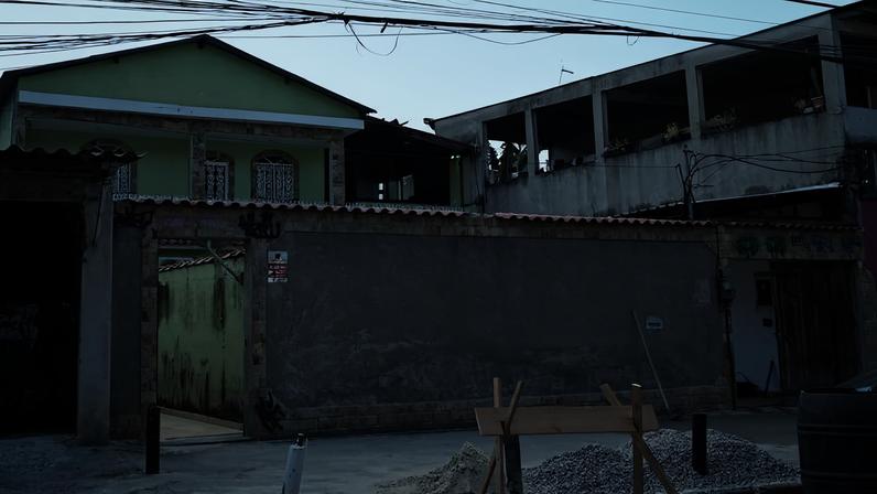 Residência verde com muro cinza alto e portão aberto à esquerda. Na frente, pilhas de areia e brita com suporte de madeira indicam obra em andamento. Fios elétricos cruzam o céu claro acima.