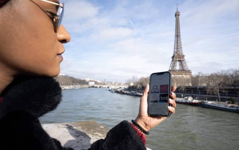 Uma mulher segura seu celular com o aplicativo de aluguel de casas de temporada Airbnb aberto na tela enquanto observa a Torre Eiffel e o Rio Sena no centro de Paris