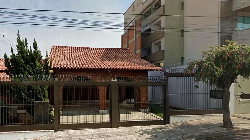 Casa térrea com telhado de cerâmica vermelha, protegida por cerca metálica com arame farpado no topo. À direita, árvore pequena na calçada e prédio residencial alto ao fundo.