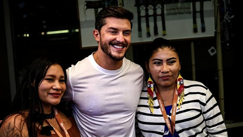 Homem de camiseta branca sorri entre duas mulheres indígenas com crachás, cercados por artesanatos coloridos pendurados.