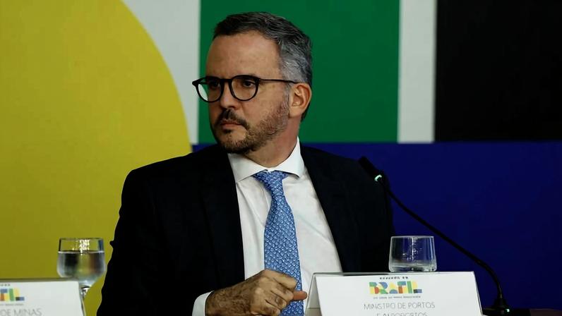 Homem de terno azul escuro, camisa branca e gravata azul clara sentado em mesa com microfone e copos de água. Fundo colorido com formas geométricas verde, amarelo e azul.