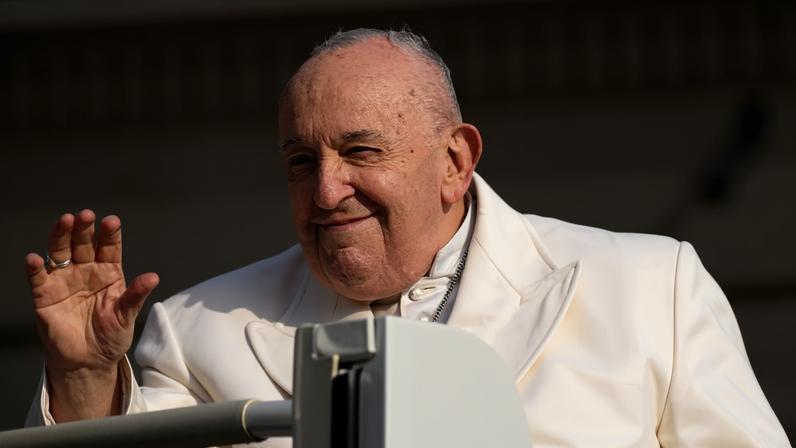 Homem idoso com vestes brancas, incluindo casula e solidéu, acena com a mão direita em gesto de saudação. Ele está em um veículo aberto, com fundo desfocado que sugere ambiente externo.
